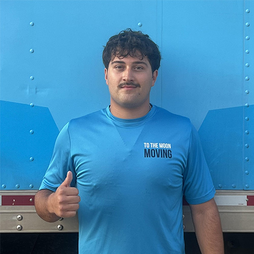 Adrian, a mover with To The Moon Moving, in a blue shirt standing next to a moving truck on a sunny day.